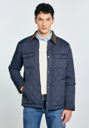 Tmavě modrá vatelínová bunda s hnědým manšestrovým límcem, dvěma náprsními kapsami a knoflíky, vrstvená přes krémový svetr s copánkovým vzorem a džínovinu.