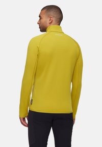 Geel fleece jack met een hoge kraag en contrasterende stiksels, voorzien van een logo op de achterkant en een getailleerd ontwerp.