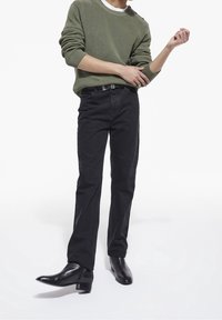 Jeans slim fit scuri con una texture liscia, abbinati a un maglione verde oliva a coste e stivali neri in pelle a punta.