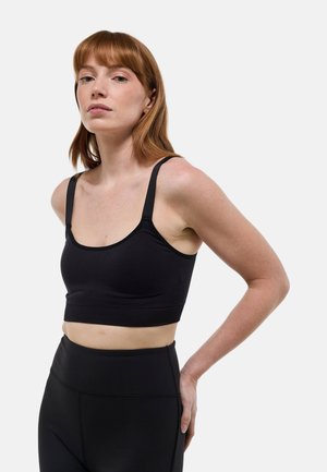 Femme aux cheveux roux portant un soutien-gorge de sport noir et des leggings noirs taille haute, debout avec une main derrière le dos.