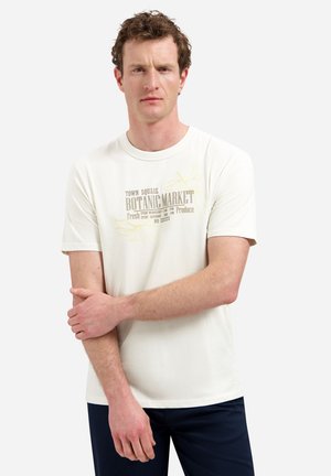 Man die een wit shirt met korte mouwen draagt met de tekst "Botanic Market", staand met gekruiste armen tegen een effen lichte achtergrond.