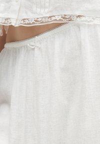 Λευκή λινή φούστα με ελαστική μέση και λεπτή δαντέλα από αντίστοιχο τοπ, που δείχνει την κοιλιά σε κοντινό πλάνο.