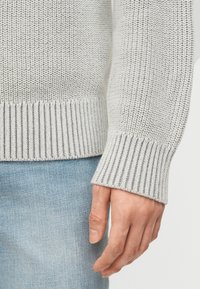 Κοντινό ενός χεριού δίπλα σε μανικετόκουμπο ελαφρώς γκρι πλεκτή μπλούζα και ανοιχτό μπλε τζιν παντελόνι.