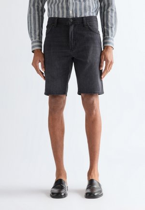 Mężczyzna ubrany w czarne szorty jeansowe, pasiastą koszulę z długim rękawem i czarne buty wsuwane, stojący na białym tle.