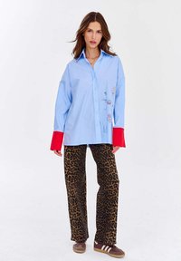 Kobieta w oversize'owej jasnoniebieskiej koszuli z czerwonymi mankietami, w spodniach w leopardzi wzór i brązowych sneakersach, stojąca na tle białym.