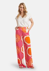 Weite Hose aus einem leuchtend pinken Stoff mit großen orangefarbenen und weißen Zitrusmustern. Kombiniert mit einem kurzärmeligen weißen Oberteil.