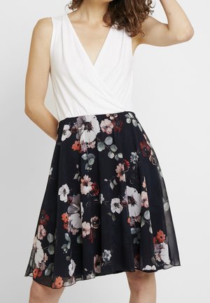 Abito senza maniche con top a incrocio a scollo a V bianco e gonna nera con motivo di grandi fiori rosa, rossi e grigi e foglie verdi.