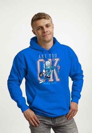 Uomo che indossa una felpa con cappuccio blu brillante con giocatore di hockey in stile cartone animato e testo "Stai bene? Spirito Puffo", in piedi davanti a uno sfondo semplice.