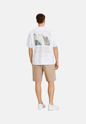 Man met wit T-shirt met cactusprint en tekst 'Azores Portugal', beige korte broek en witte sneakers, kijkend van achteren op een witte achtergrond.