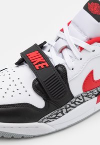 Zapatilla Nike Air Jordan con parte superior blanca, detalles en cuero negro, logo rojo, mediopie en gris texturizado y correa ajustable.