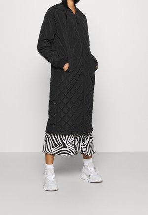 Person iført en lang sort quiltet frakke over et sort-hvidt zebramønstret skørt, hvide sokker og hvide sneakers, stående mod en ensfarvet baggrund.