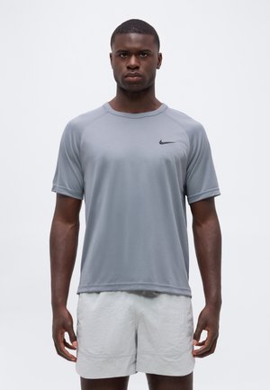 Mand stående vendt fremad iført en lysegrå Nike kortærmet skjorte og hvide shorts mod en ensfarvet baggrund.