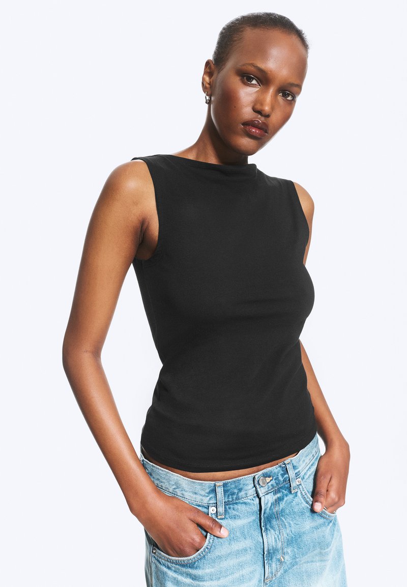Femme aux cheveux courts portant un haut noir sans manches et un jean bleu clair, debout les mains dans les poches devant un fond uni.
