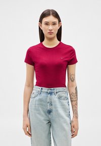 Ung kvinde med langt mørkt hår iført en tætsiddende rød t-shirt og lyseblå højtaljede jeans, der viser tatoveringer på sin venstre arm.
