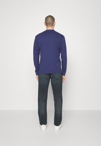 Mörkblå skjorta med långa ärmar, i bomullsmaterial, platt krage, förstärkta sömmar. Bärs med mörka jeans och vita sneakers; sedd bakifrån.