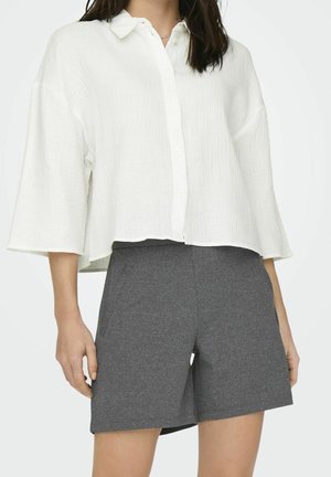 Mujer con una camisa blanca suelta abotonada con mangas anchas y pantalones cortos oscuros de longitud hasta la rodilla sobre un fondo liso.