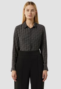 Schwarzes, gemustertes Hemd mit kragenloser Halslinie, das ein dezentes Wirbelmuster in heller Farbe aufweist, kombiniert mit hoch geschnittenen schwarzen Hosen.