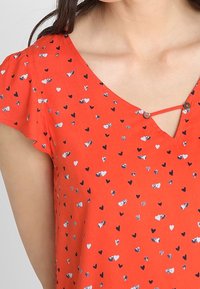 Rode blouse met een V-hals, korte gerimpelde mouwen en een knoopdetail bij de halslijn. Kenmerkt zich door een verspreid hartpatroon in verschillende kleuren.