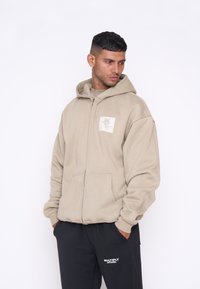 Beige mikina na zip s předními kapsami a nášivkou. Má uvolněný střih a kapuci se šňůrkou. Nošena s černými kalhotami a minimálními detaily.