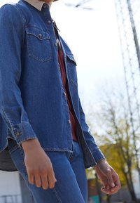 Persona vestita con giacca di jeans blu e jeans, in piedi all'aperto vicino ad alberi e a una struttura metallica alta in una giornata di sole.