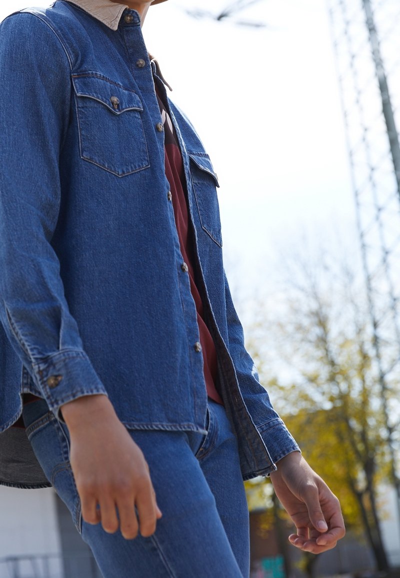 Persona vestita con giacca di jeans blu e jeans, in piedi all'aperto vicino ad alberi e a una struttura metallica alta in una giornata di sole.