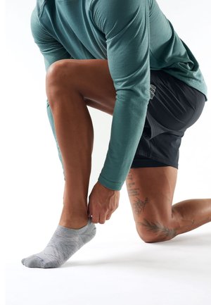 Person kniet auf einem Knie, passt eine graue Knöchelsocke an, trägt ein türkises Langarmshirt und schwarze Shorts, mit sichtbaren Tattoos am Bein.