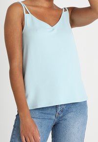 Ljusblå ärmlös topp med V-ringning och tunna axelband, som visar en texturerad tyg och avslappnad passform. Kombinerad med jeans i denim.