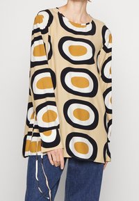 Långärmad blus i beige med stora svarta och senapsgula ovala mönster. Har en rund halsringning och lös passform. Mjuk, slät tyg.