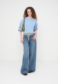 Pull tricoté bleu clair avec des détails floraux brodés sur de larges manches, associé à un jean bleu taille haute à coupe ample et un sac vert.