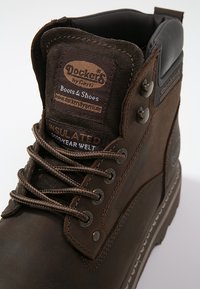 Botas de trabajo aisladas marrones con parte superior de cuero, cuello acolchado, ojales de metal y costura Goodyear. Presenta un logo y costuras en contraste.