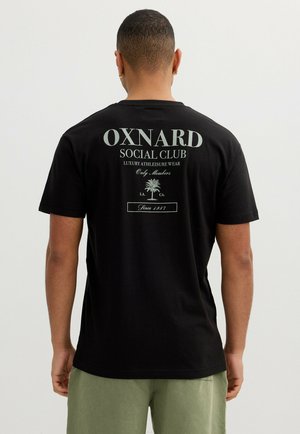 Hombre de espaldas vistiendo una camiseta negra con el texto "OXNARD SOCIAL CLUB" y un gráfico de palmera, combinado con pantalones cortos verde claro.