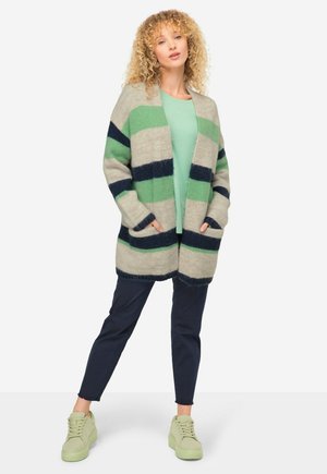 Cardigan lavorato a maglia con strisce verde e blu navy, apertura frontale, due tasche, indossato sopra una maglietta verde chiaro, abbinato a pantaloni blu navy e sneakers verde chiaro.