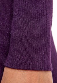 Tessuto a maglia ribattuto viola con una lucentezza metallica. Il primo piano mette in risalto una superficie testurizzata e un’apertura della manica aderente.