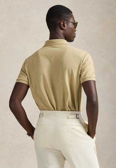 Άνδρας γυρισμένος με την πλάτη, φοράει ανοιχτοκίτρινη πόλο μπλούζα, εκρού παντελόνι με ρυθμιστές στο πλάι, γυαλιά ηλίου και ρολόι χειρός, σε ουδέτερο φόντο.