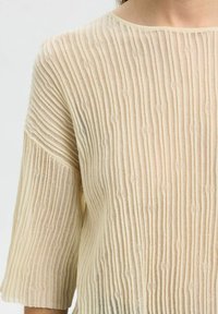 Hellbeige gestricktes Oberteil mit vertikal gerippter Textur, kurzen Ärmeln und rundem Ausschnitt. Das halbtransparente Material erzeugt einen gestuften Look.