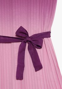 Lila tygsrosett knuten runt midjan på en plisserad rosa klänning, som framhäver de texturerade vertikala linjerna och färgkontrasten.