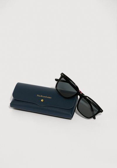 Ochelari de soare negri cu lentile întunecate și rame pătrate, lângă un etui din piele bleumarin cu litere din aur. Etuiul are un capac rotunjit.
