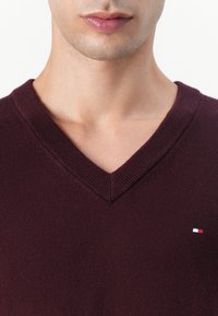 Burgundi színű kötött pulóver V-nyakkal, bordázott részletekkel és kis logóval a bal mellen. Puha tapintású.
