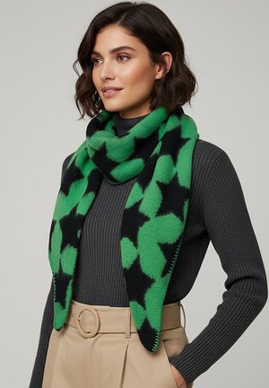 Woman wearing a green and black star-patterned scarf over a dark grey ribbed sweater and beige high-waisted pants with a round belt buckle.