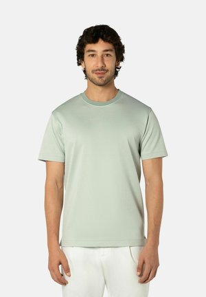 Man met donker krullend haar, gekleed in een lichtgroen shirt met korte mouwen en witte broek, staand tegen een effen lichte achtergrond.