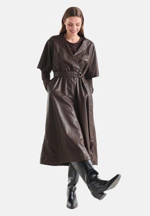 Vrouw draagt een donkerbruine leren jurk met riem over een kanten top, zwarte kniehoge laarzen, staat met één been lichtjes opgetild en handen in de zakken.