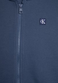 Mörkblå zip-up hoodie tillverkad av mjukt tyg, med en central dragkedja och en liten, broderad logotyp på vänster bröst.