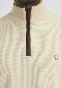 Pull crème en tricot avec une fermeture éclair marron, présentant un motif texturé et un petit logo sur le coin inférieur droit.