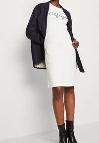 White fitted dress with handwritten text in navy. Layered with a dark navy blazer. Black ankle boots. Smooth fabric texture. Simple design.