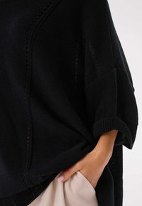 Czarny dziany ponczo o luźnym kroju, szerokich rękawach i teksturowanej tkaninie. Posiada żebrowany dół i boczne detale. Ręka spoczywa w kieszeni.