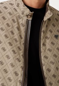 Giacca beige con motivo a monogramma marrone scuro su tutta la superficie, zip marrone, colletto con bottoni e maglietta nera visibile sotto.