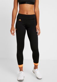 Kvinna i svarta cropped leggings med orange accenter och vita Adidas sneakers, stående mot en enkel bakgrund.