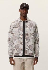 Ζακέτα πατςγουορκ σε αποχρώσεις του μπεζ και του γκρι, με ποικίλες υφές, διακριτικά μοτίβα και ελαφριά κουμπιά, φορεμένη πάνω από ένα σκούρο πουκάμισο.