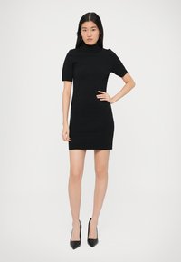 Crna pletena haljina s visokim ovratnikom, kratkim rukavima i uskom siluetom. Nošena s crnim štiklama sa špicastim vrhom. Glatka tekstura, minimalni uzorak.