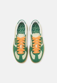 Un paio di sneaker in suede verde e beige con lacci arancioni e suole interne verdi a motivo Puma, viste dall'alto su uno sfondo bianco.